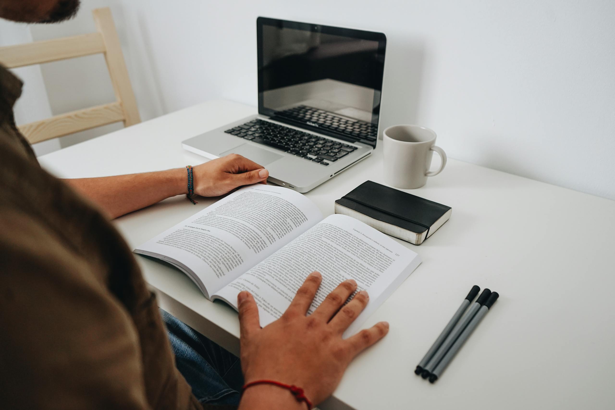 A person reading at a minimalist home office with a laptop, coffee mug, and book.